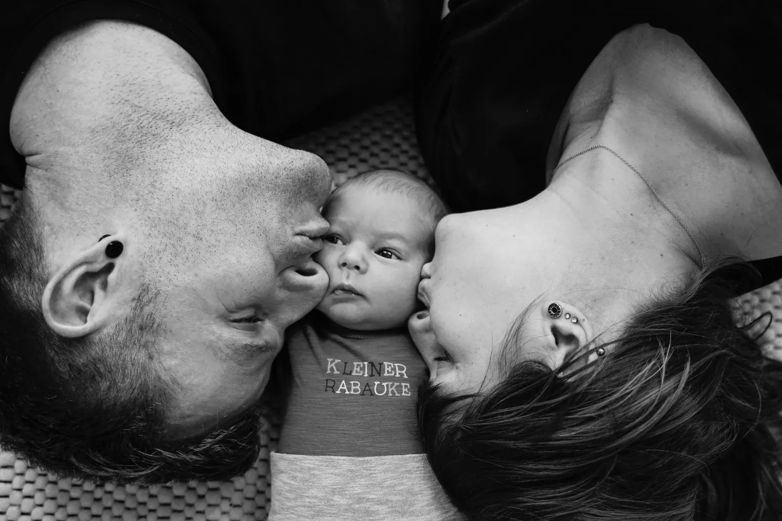 Schwarzweißbild: Ein Baby liegt zwischen den Gesichtern seiner Eltern
