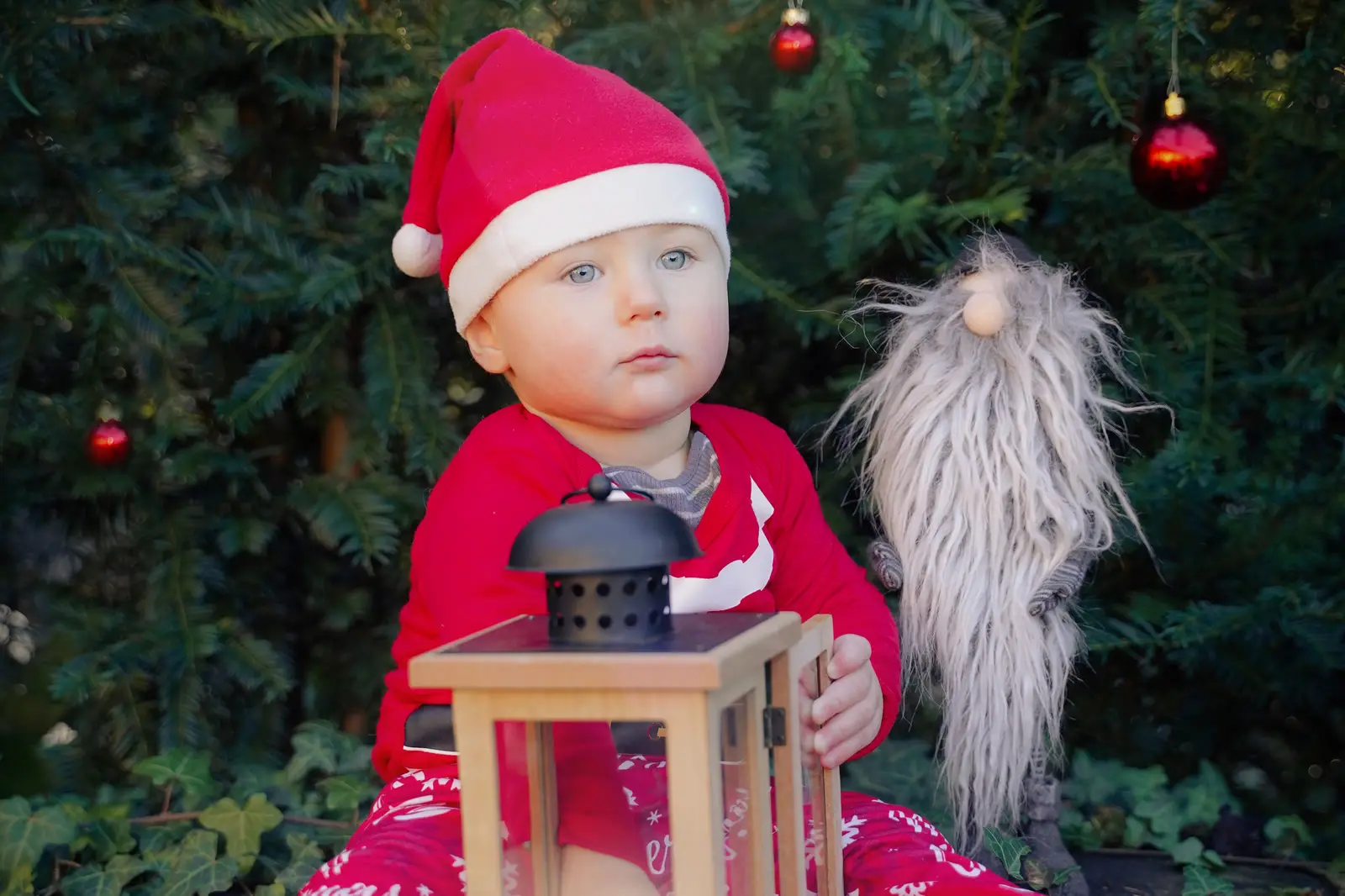 Ein Kleinkind mit roter Weihnachtsmütze sitzt neben einer leuchtenden Laterne vor einem Tannenbaum