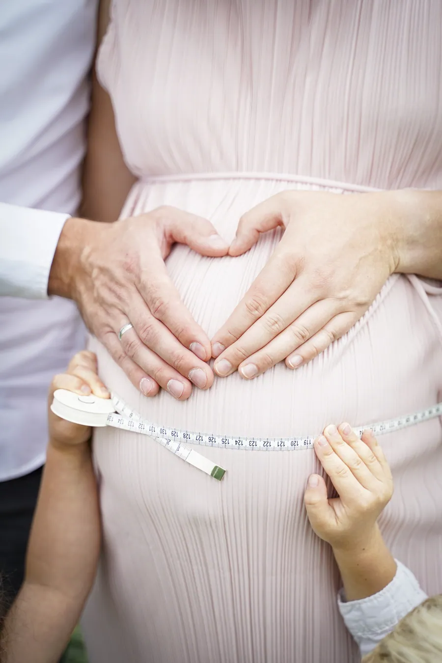 Zwei Hände formen ein Herz auf dem Babybauch, eine Kinderhand hält ein Maßband daneben