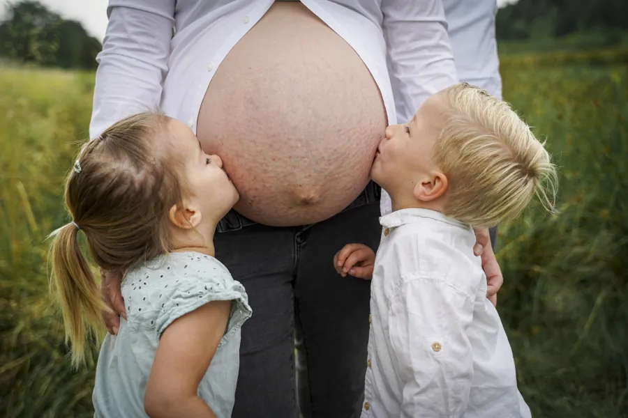 Zwei Kinder küssen von beiden Seiten den Babybauch ihrer Mutter auf einer Wiese