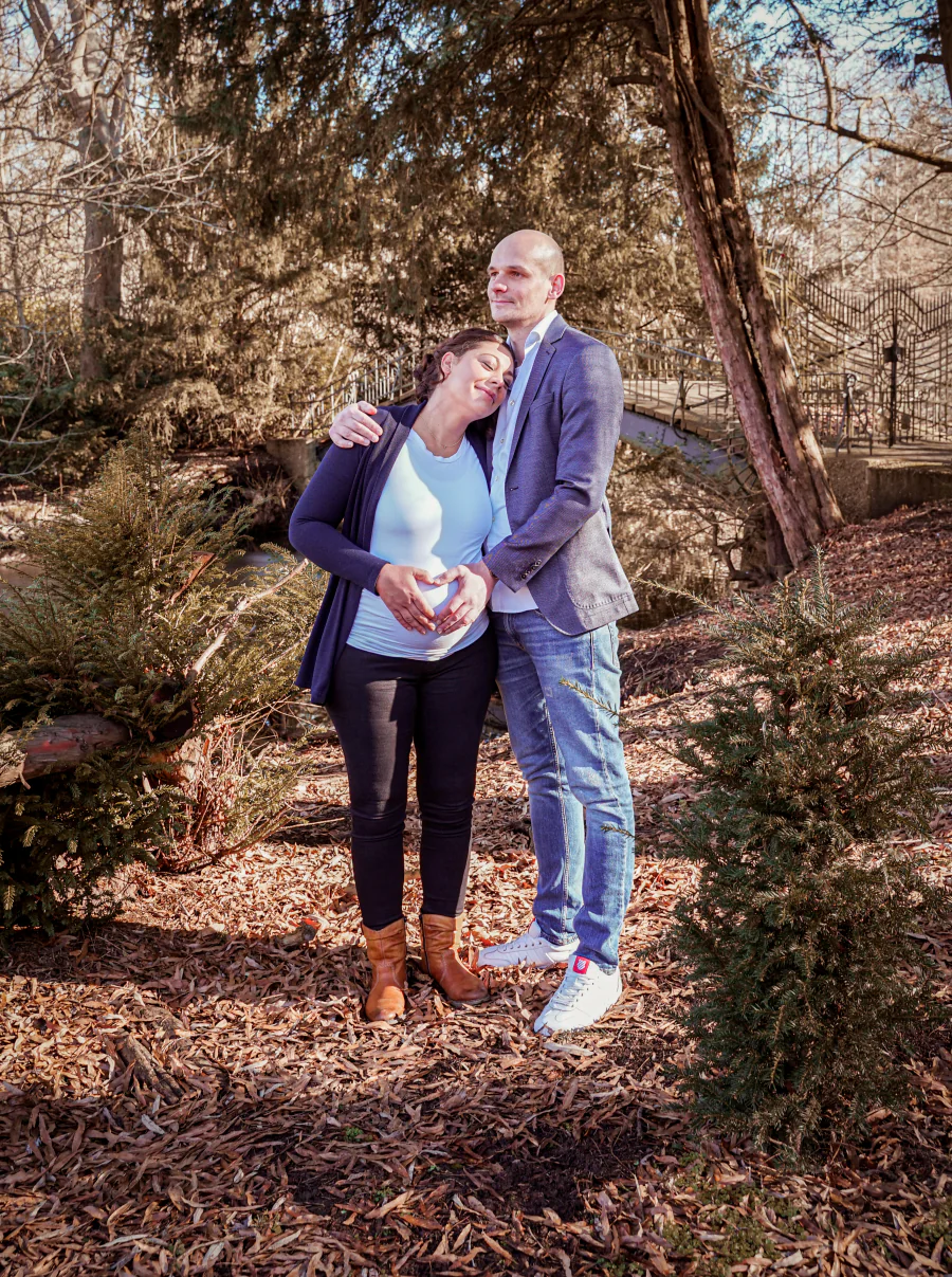 Eine schwangere Frau steht mit einem Mann in einem herbstlichen Wald, beide halten die Hände an ihren Bauch