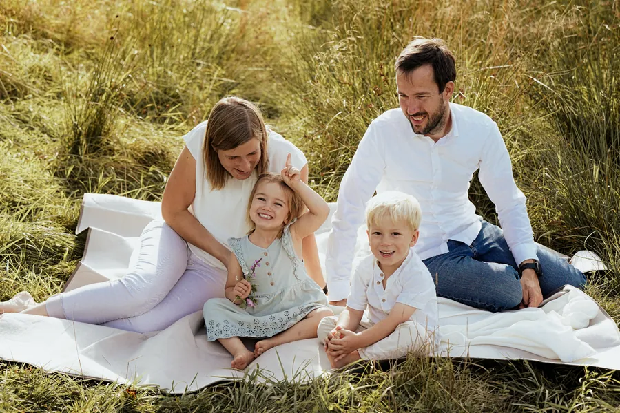 Eine vierköpfige Familie sitzt lachend auf einer Picknickdecke im hohen Gras