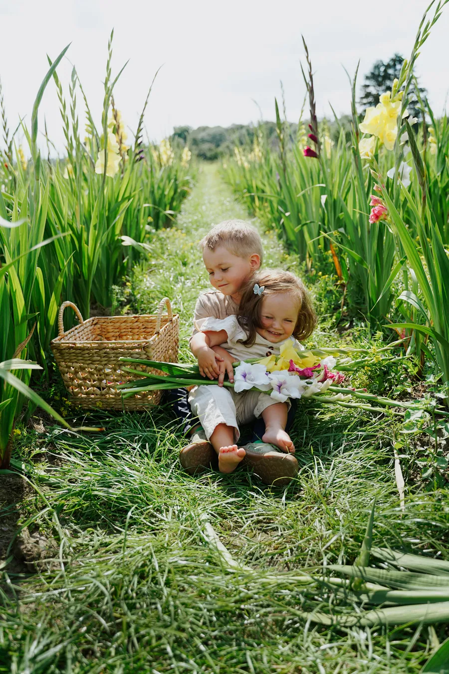 Zwei kleine Kinder sitzen lachend auf einem Weg zwischen hohen Gladiolen, daneben ein Korb voller Blumen