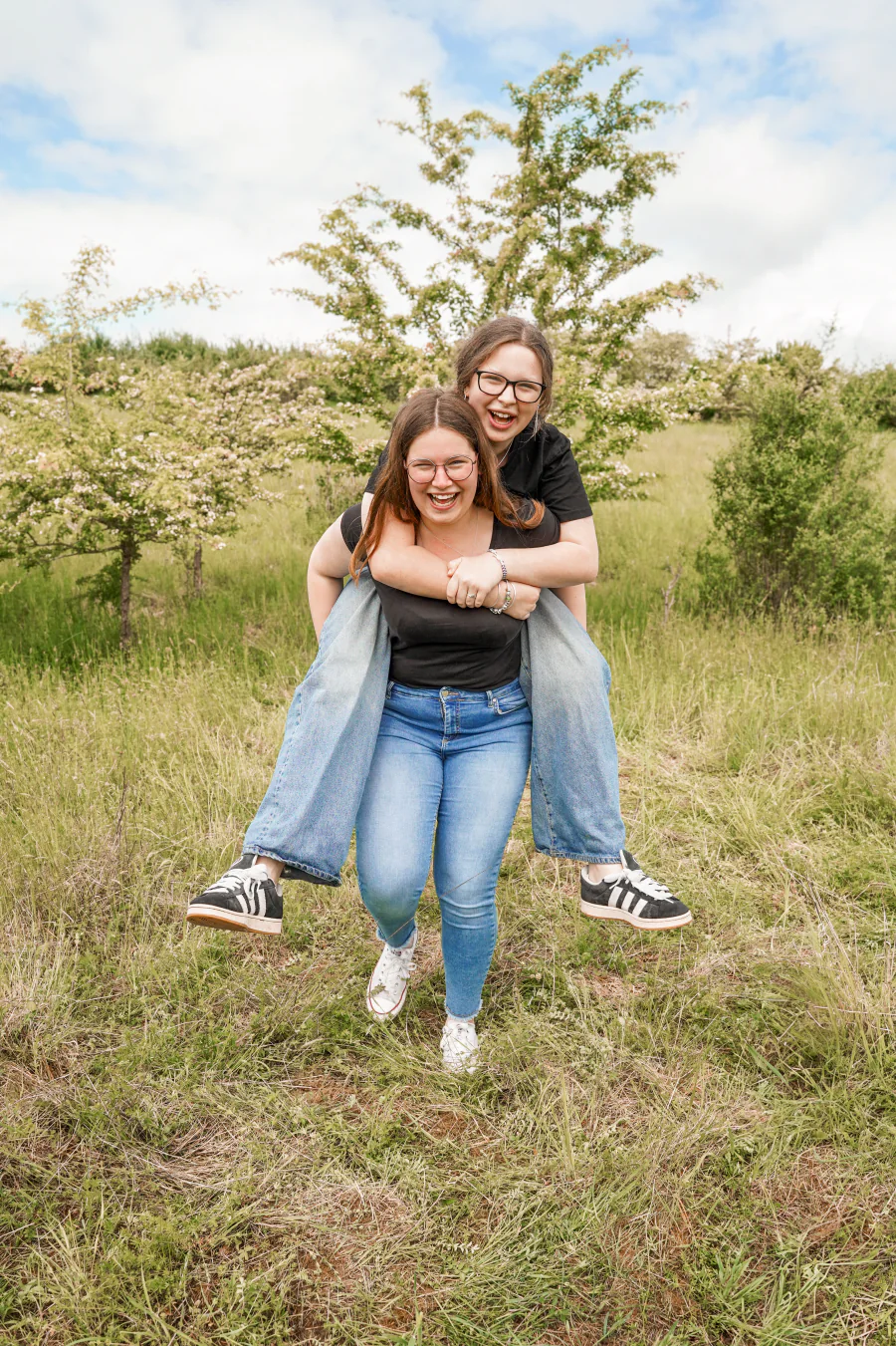 Zwei Frauen lachen auf einer Wiese, eine trägt die andere huckepack