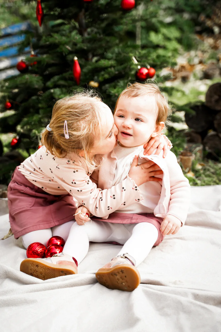 Ein kleines Kind küsst ein lachendes Baby vor einem Weihnachtsbaum