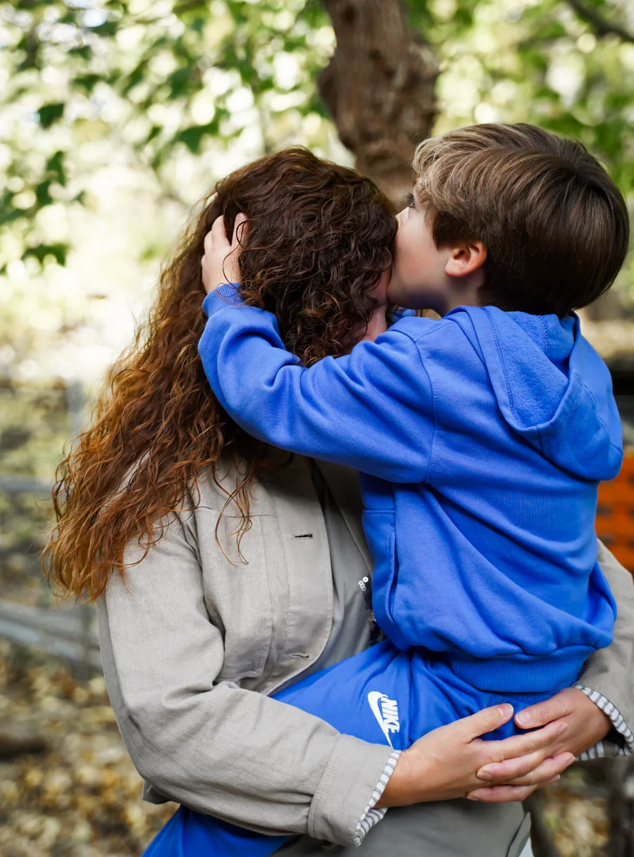 Ein Kind im blauen Pullover umarmt eine Frau zwischen herbstlichen Bäumen