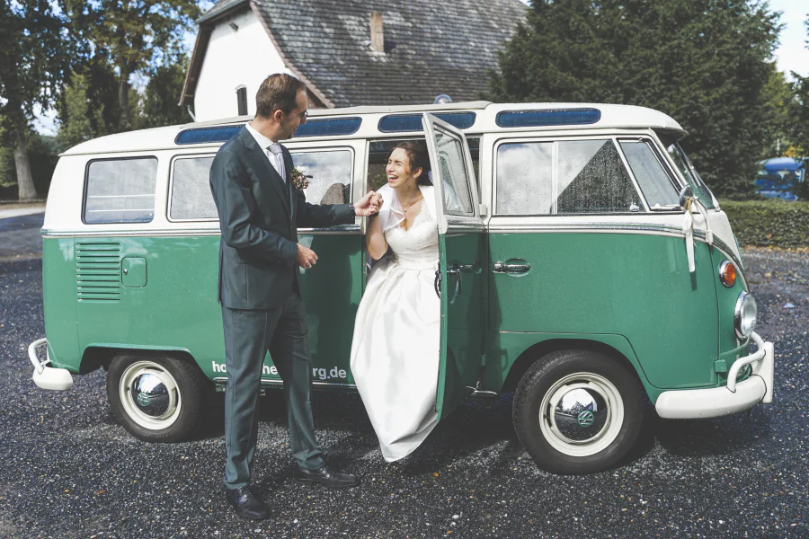 Ein Brautpaar steht lachend vor einem grün-weißen VW-Bus