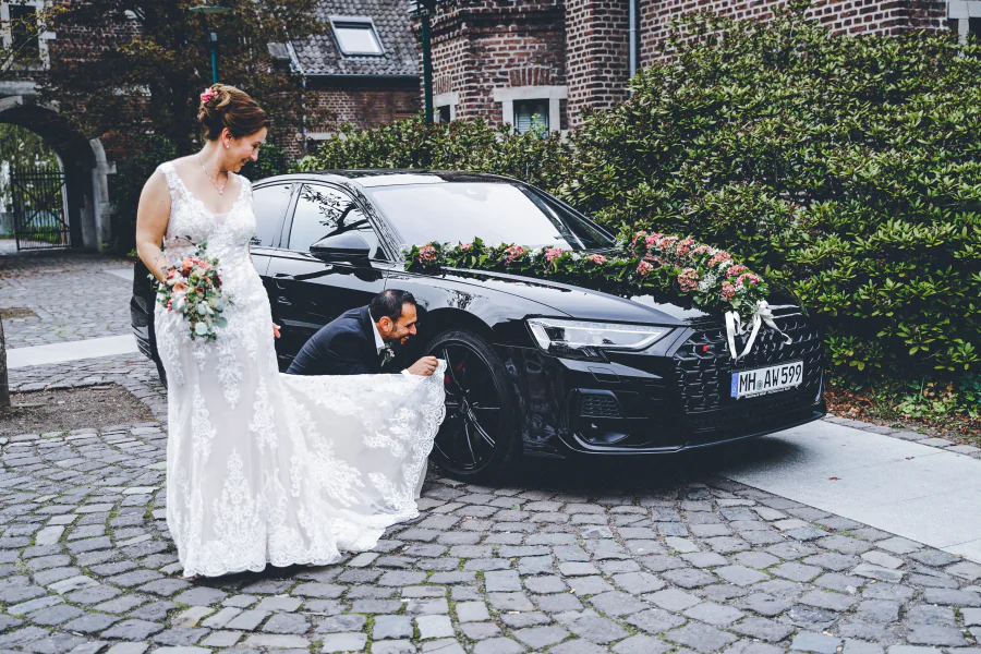 Eine Braut steht mit ihrem Blumenstrauß neben einem geschmückten schwarzen Auto
