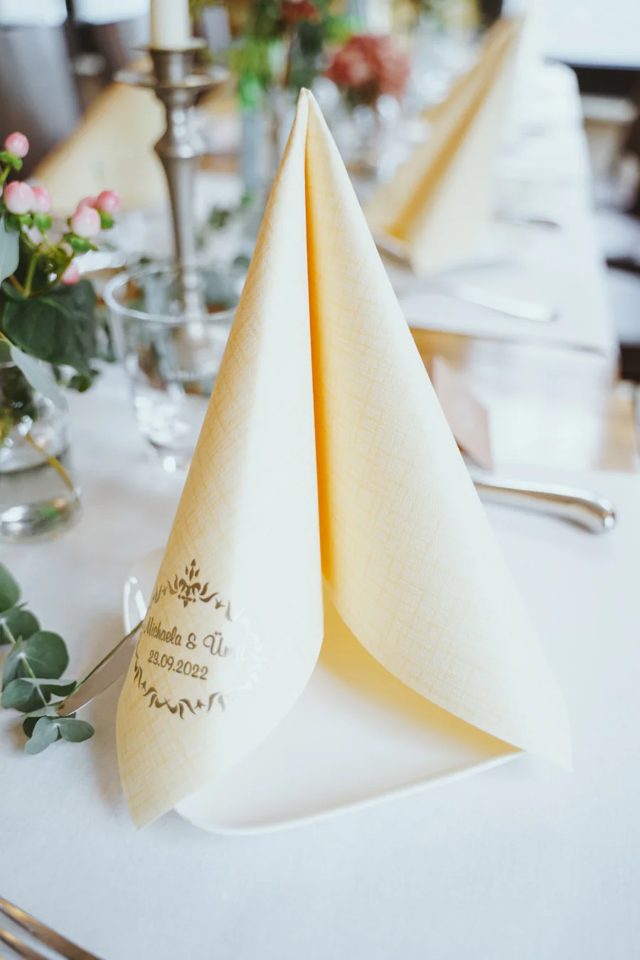 Eine gefaltete Stoffserviette mit handschriftlicher Beschriftung steht auf einer Hochzeitstafel