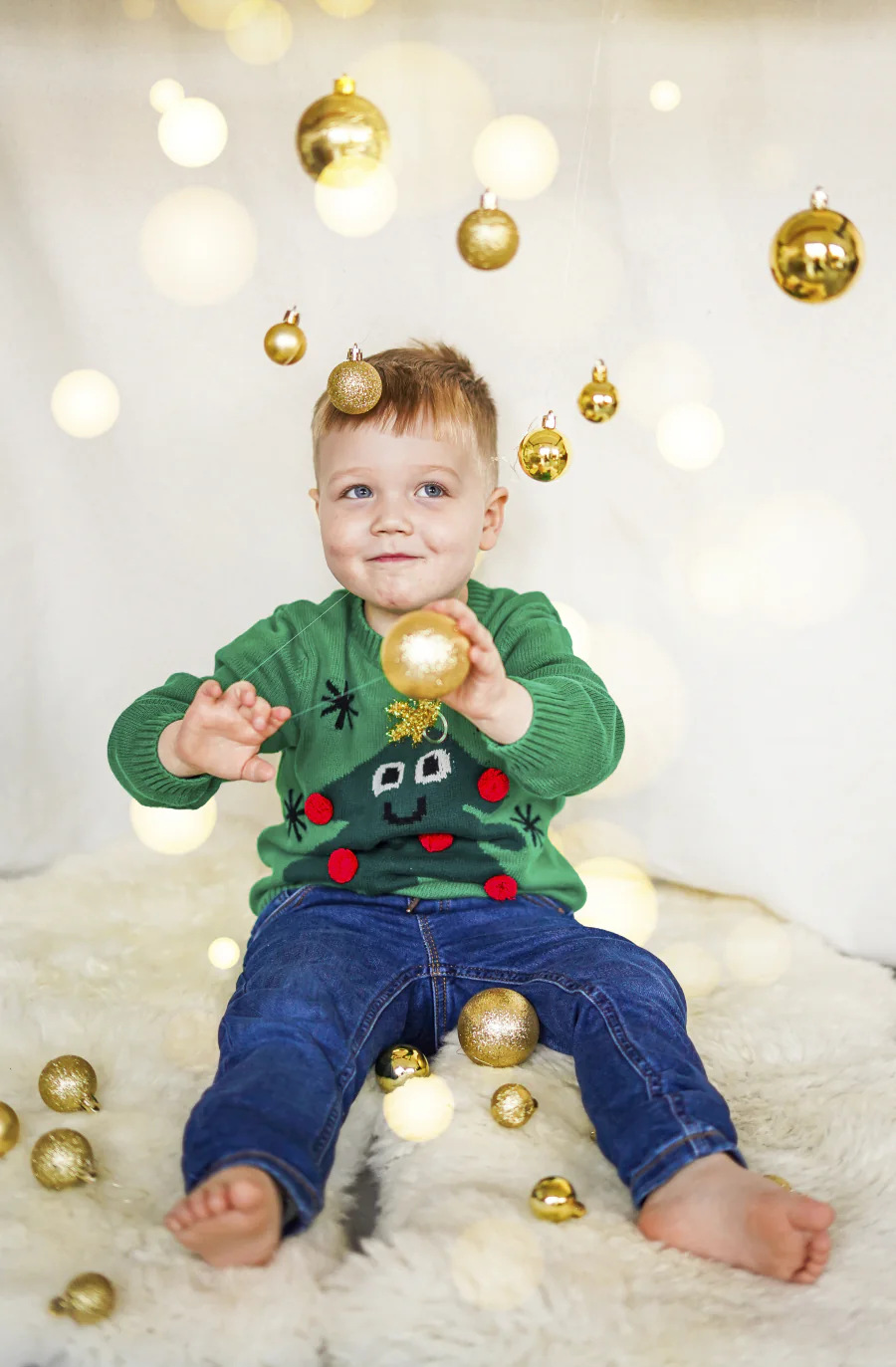 Ein Kleinkind sitzt zwischen goldenen Weihnachtskugeln vor einer hellen Lichterkulisse