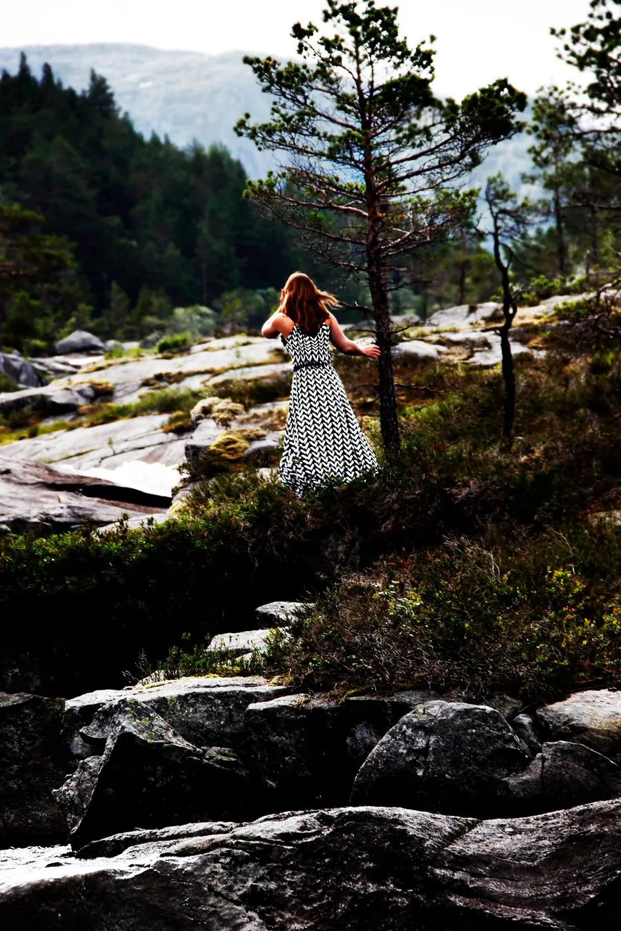 Eine Frau im gemusterten Kleid steht in einer felsigen Berglandschaft an einer Kiefer