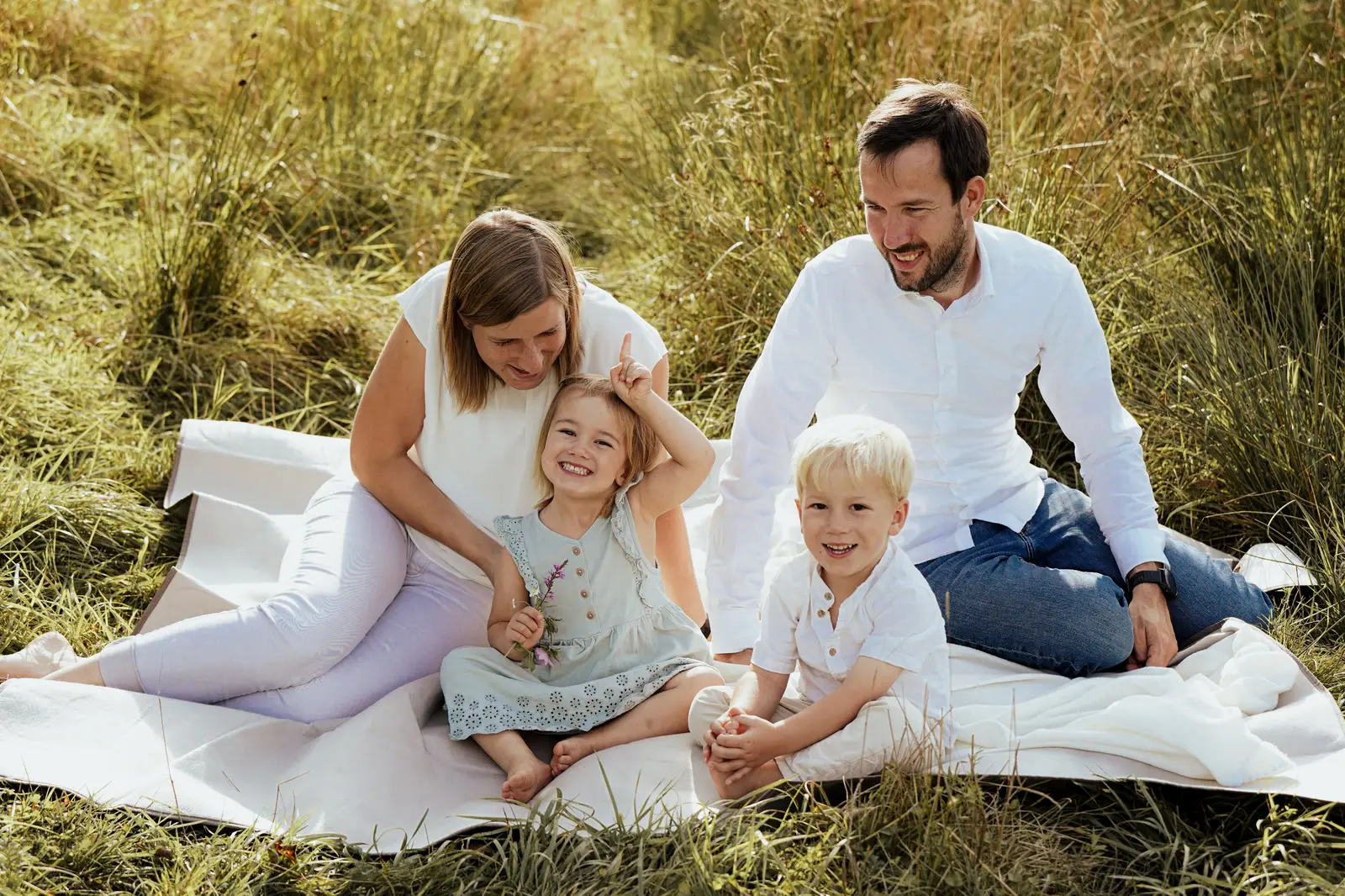 Zwei Erwachsene und zwei Kinder sitzen lachend auf einer Decke im hohen Gras.