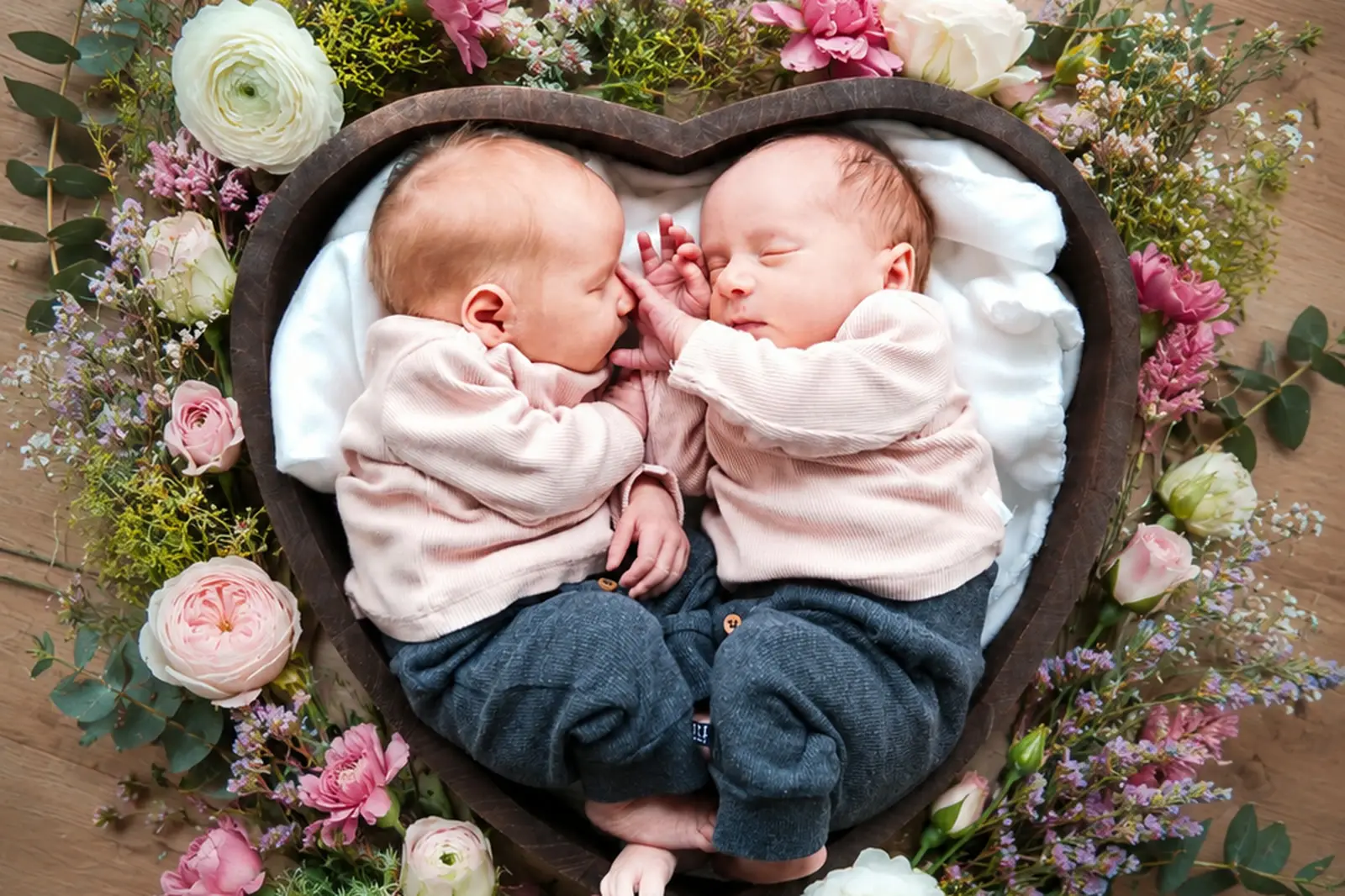 Zwei schlafende Babys liegen in einem herzförmigen Holzbett zwischen Blumen